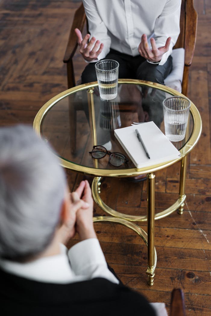 about-us Two adults discussing mental health in a counseling session across a glass table indoors.